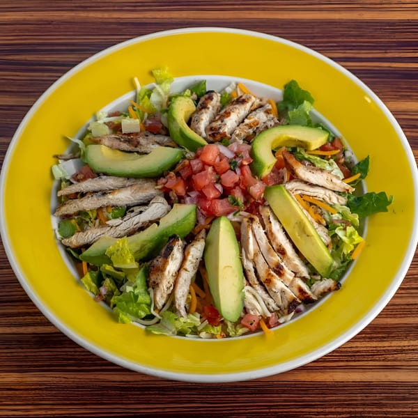 Grilled chicken and avocado salad with pico de gallo, lettuce, and shredded cheese in a yellow bowl.