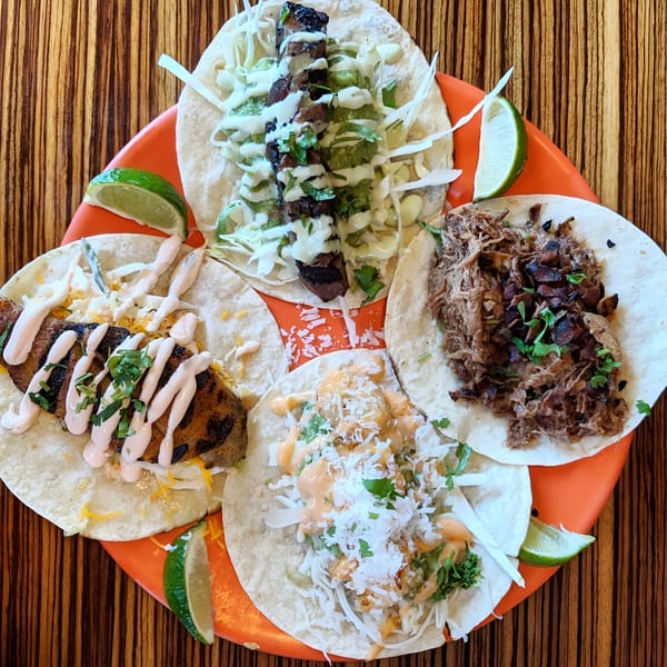 Four Mexicali Blue tacos with various fillings and sauces, garnished with lime wedges on an orange plate.