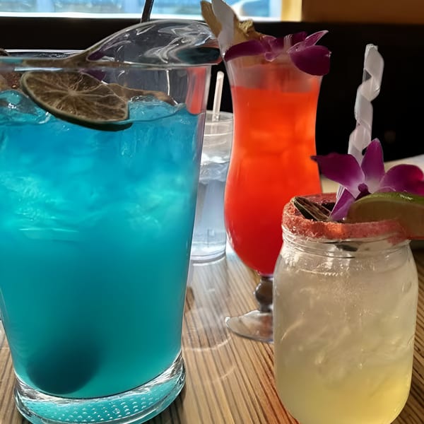 Three colorful Mexicali Blue cocktails, including blue, red, and yellow drinks, on a wooden table.
