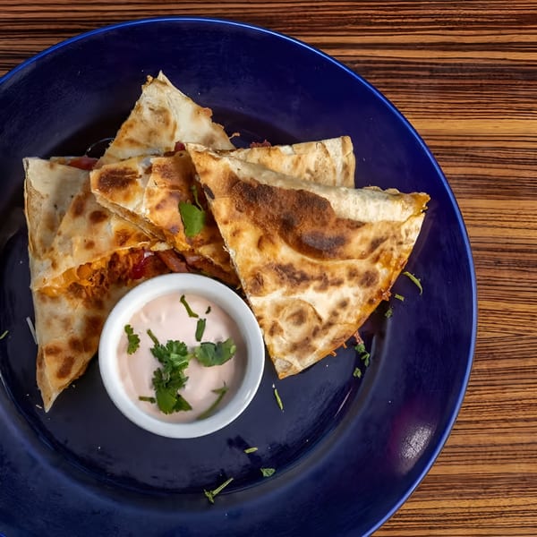 Four quesadilla wedges with a side of creamy sauce, garnished with cilantro on a blue plate.